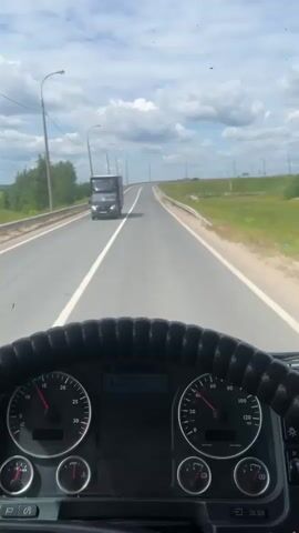A first-person view from the driver's seat of a truck shows the road ahead and the truck's dashboard. The road is paved with white lane markings and is bordered by a guardrail and green fields with trees in the distance. The sky is blue with scattered clouds. On the dashboard, the speedometer indicates the truck is traveling at approximately 40 mph. A black truck is visible ahead in the left lane. The video also includes a brief shot of the side mirror and window.