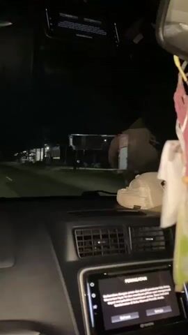 The video shows the interior of a car at night, looking out the windshield. The dashboard is visible in the foreground, with a car's infotainment system displaying a warning message in Indonesian. To the right, a rearview mirror hangs from the windshield, decorated with a colorful macrame charm. Outside the car, a dark, unlit road is visible, bordered by buildings and sparse streetlights. The car appears to be in motion, evidenced by the slight blur and the receding perspective of the road. No people are directly visible within the car, but a shadowy figure can be seen through the windshield in the distance. The overall atmosphere is dark and quiet, suggesting a nighttime drive through a semi-urban or rural area.