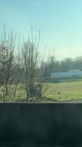 The video shows a view from a moving vehicle, likely a train or car, passing a green field. In the background, there are trees and a tall electricity pylon. There is a small green structure on the right edge of the field. The sky is a pale blue. The view is slightly obscured by a concrete barrier or wall at the bottom of the frame.
