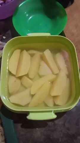 The video shows a close-up, overhead view of a light green, rectangular plastic container filled with water and potato wedges. The potatoes are cut into irregular wedges, some thicker than others, and appear to be raw. The container is filled with enough water to cover the potatoes. In the background, there is a green plastic bowl, also open, and a hint of a purple container. The lighting suggests an indoor setting. The camera perspective is static, looking directly down into the container. No people or hands are clearly visible interacting with the potatoes or container. The overall impression is of food preparation, specifically the pre-treatment of potatoes in water.