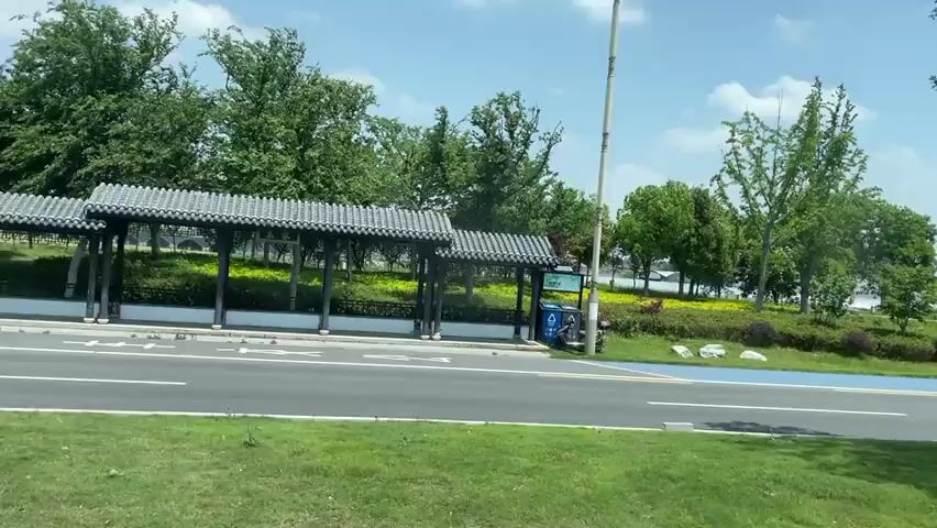 The camera, seemingly mounted in a moving vehicle, captures a view of a well-maintained roadside landscape on a bright, sunny day. The road is marked with white lines and a blue lane on the shoulder. Lush green grass covers the verge, dotted with bushes and trees. In the initial shot, there is a small bus stop structure with a tiled roof and a scooter parked beside it. As the vehicle moves forward, the view expands to reveal more trees, landscaping, rocks, and a crosswalk. The sky is blue with some scattered clouds. In the distance, there appear to be buildings or structures.