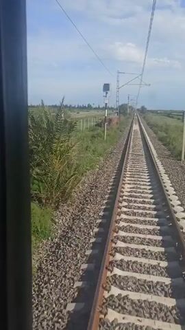 The video captures a train ride on a bright, sunny day, with the camera positioned inside the train, looking out. The primary focus is the railway track stretching straight ahead into the distance, flanked by gravel. On either side of the track, there is green vegetation, including grass and some shrubs. A road runs parallel to the tracks on the left side, with a car visible moving in the distance. Overhead, there are overhead power lines supported by metal poles. The sky is blue with some scattered white clouds. The camera is static, creating a sense of motion as the train progresses forward.