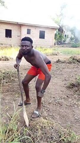 A man stands in a field, leaning on a shovel and making a face at the camera. He is shirtless and wearing orange shorts and slides. He has a wrist watch on his left wrist. A cinderblock house stands in the background. The ground is mostly dirt with patches of grass.