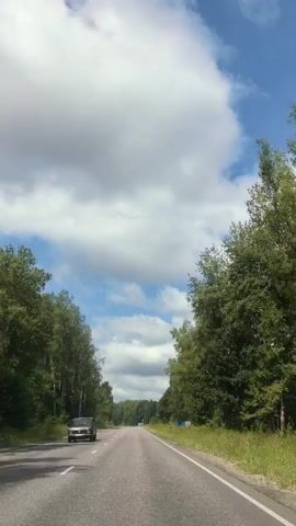 A first-person perspective video shows a car driving down a two-lane asphalt road. The road is lined with tall green trees on both sides, creating a tunnel-like effect. The sky above is a bright blue with scattered white clouds. A few other cars are visible in the distance. The car the video is being filmed from passes a dark-colored vintage car on the left side of the road.