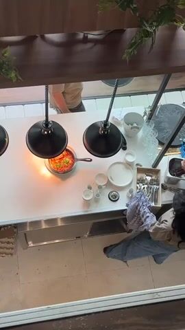 The video shows an overhead view of a kitchen counter. There are two people visible. On the left, a person wearing a black t-shirt is placing a tray of food onto the counter. On the right, a person wearing a beige top and blue jeans is holding a patterned cloth and appears to be wiping down the counter or arranging utensils. In the center of the counter, a pan of food is cooking under a heat lamp. There are several plates, bowls, and cups arranged on the counter, as well as a bucket of ice and glassware. The environment appears to be a professional kitchen or food service area.