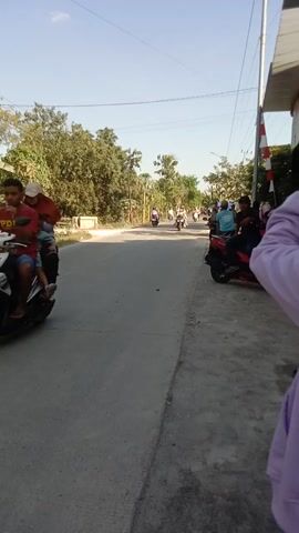 A person wearing a light purple hooded sweatshirt is partially visible in the foreground, facing away from the camera. The scene is outdoors, with a clear blue sky. A paved road runs diagonally across the frame. Several motorcycles are on the road, some with riders. On the left side of the road, there are trees and vegetation. A white pole with red and white striped markings is visible near the top left of the frame. Another person, wearing a blue head covering and a blue shirt, is visible further down the road, also on a motorcycle. The camera is tilted sideways.