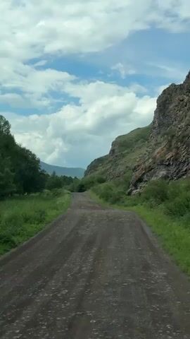 The video shows a perspective from a moving vehicle on a gravel road. To the right of the road is a steep, rocky incline covered in green vegetation. To the left of the road is a field of green grass and trees. The sky is blue with scattered white clouds. The road curves ahead and disappears into the distance. The overall environment appears to be a natural, mountainous landscape.