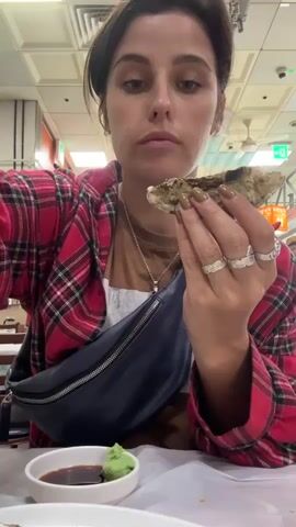 A woman in a red and black plaid shirt over a brown t-shirt and a dark blue fanny pack around her waist, is sitting at a table. She is holding an oyster with a silver fork and knife. In front of her is a small white dish containing soy sauce and a dollop of green wasabi. The woman brings the oyster to her mouth and eats it. The background appears to be a restaurant with fluorescent lighting and some signs.
