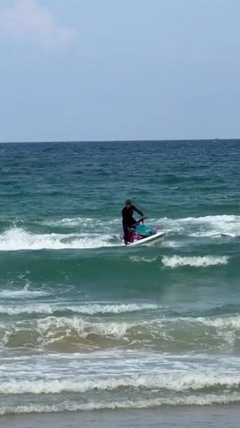 The video shows a person on a jet ski on the ocean, not far from a sandy beach. The person is wearing a black long-sleeved shirt and hat. The jet ski is primarily white with purple and turquoise accents. The waves are small to medium-sized, and the person rides the jet ski over them. The ocean is a turquoise color and the sky is blue.