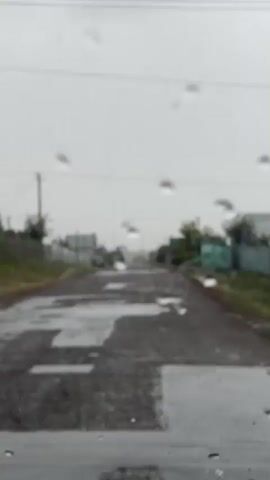 The video shows a view through a wet windshield, likely from inside a vehicle, on a cloudy, possibly rainy day. Water droplets are visible on the glass, obscuring the view to some extent. Outside, a paved road stretches into the distance, with puddles of water indicating wet conditions. Along the sides of the road, there are houses and fences, suggesting a rural or suburban setting. Trees and other vegetation are also present. The overall atmosphere is overcast and somewhat bleak, consistent with rainy weather. The vehicle appears to be moving, as the perspective shifts slightly throughout the short clip. There are no people or animals clearly visible.