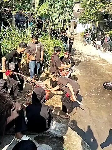 A group of people are gathered outdoors, seemingly in a village setting. There is a central area where several individuals are engaged in some form of ritual or performance. Some are dressed in traditional attire, including black clothing and headbands. One person appears to be lying down, possibly part of the performance, while others surround them. There are onlookers standing and sitting around the periphery. In the background, there are houses, trees, and lush vegetation. The ground appears to be a mix of dirt and paved surfaces. The overall atmosphere suggests a cultural event or gathering.