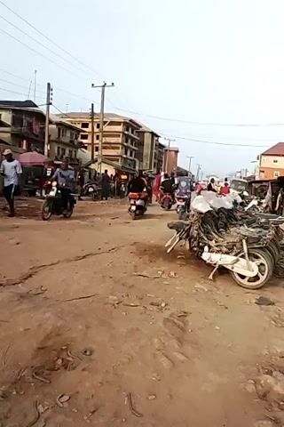 The video captures a dynamic street scene in Nnewi, Nigeria, during what appears to be daytime under an overcast sky. The unpaved, dusty road is central, marked with tire tracks and debris.

In the foreground to the right, a large pile of dismantled or damaged motorcycle parts, including wheels and frames, lies on the ground, with a red plastic crate positioned beside it. Throughout the scene, numerous people are present, with some walking and others gathered around market stalls or buildings. Notably, a group of individuals wearing red shirts is seen moving rapidly across the street from left to right around the 0:02-second mark.

Motorcycles are a prevalent feature, with several being ridden along the street and others parked or stationary. Along the left side of the road, various market stalls or shelters with umbrellas are set up, indicating commercial activity. Buildings, some multi-story, line both sides of the street, defining the urban landscape. Overhead power lines strung between poles crisscross the sky. The overall atmosphere is bustling and active, characteristic of a lively town or market area. No discernible text is visible.