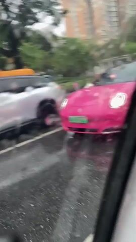 This is a first-person perspective video filmed from inside a vehicle. The view is out the side window, which is wet with raindrops. The video shows traffic on a wet road. In the foreground, a portion of the interior of the filming vehicle is visible. In the background, several cars are visible. A pink car is in the mid-ground, and a silver SUV is to its left. Further back, a white SUV is visible, and behind it are a red bus and an orange vehicle. The road is wet, suggesting it is raining or has recently rained. The overall impression is a traffic scene on a rainy day.