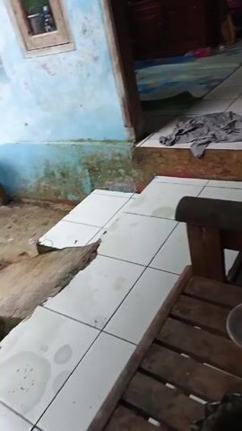 Here is a detailed description of the video:

The camera is placed in an outdoor setting, looking into what appears to be the interior of a house. The view is from a low angle. The foreground features a rustic wooden chair with a small, round, translucent lid resting on its surface. The chair is positioned on white tiled flooring that extends toward the entrance of the house.

To the left of the frame, a light blue wall shows signs of wear and tear, with areas of paint peeling or missing. The entrance to the house is visible, leading into a space where a wooden cabinet is present. In the interior, a light blue patterned sheet or tarp partially covers the floor. A gray garment is also casually laid on the tiles near the entrance.

The overall impression is of a slightly run-down or well-used area, combining both indoor and outdoor elements. There is a sense of age and possibly a lack of consistent maintenance, evident in the state of the wall and the tiled flooring. The wooden chair and the objects placed on it contribute to the scene's homely, unorganized atmosphere.