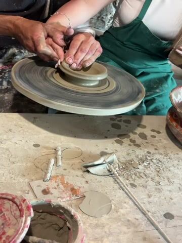 This video captures a pottery-making scene in Medovo, Bulgaria, divided into two frames.

The top frame focuses on the hands of two individuals working on a potter's wheel. A person with darker skin and visibly clay-stained hands appears to be guiding the hands of another person with lighter skin and manicured nails, who is wearing a green apron over a white top. Together, they are shaping a small, unglazed clay bowl on the rapidly spinning wheel. Both sets of hands are covered in wet clay, indicating active participation in the process.

The bottom frame shows a closer view of the workspace, revealing a messy, clay-splattered concrete or wooden surface. Various pottery tools are scattered around, including wire cutting tools, a loop tool, and what appears to be a rib tool. There are also several small pieces of dried clay and two plastic containers, one filled with wet clay slurry and the other possibly with water or a different type of clay. The overall scene suggests a hands-on, active pottery studio environment.