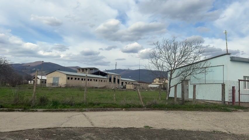 The video shows a rural scene with several low, industrial-style buildings behind a chain-link fence and a paved road. The buildings are light tan or blue. In the background, there are hills or mountains under a cloudy sky. A bare tree stands to the right of the frame, partially obscuring a building.  There's no discernible activity or people visible.