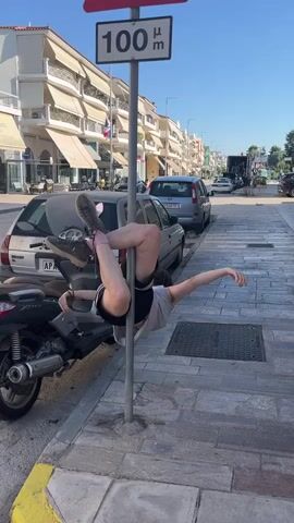 A person is performing a pole dance on a street. The person is wearing shorts and a t-shirt. They are in a contorted position, wrapped around a pole. A scooter is parked next to them. In the background, there are buildings, cars, and a street sign that reads "100 μm". The sky is blue and clear.