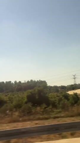 The video captures a view from a vehicle traveling along a road, likely a highway, in Shafir Regional Council, Israel. The scene is dominated by a clear, pale blue sky, indicating a bright, possibly sunny day. The landscape consists of rolling hills covered in dry, brown vegetation and interspersed with areas of green trees and shrubs. In the distance, a line of trees forms a dense foliage across the horizon.

Several man-made objects are visible, suggesting the proximity of human infrastructure. These include what appear to be power lines supported by tall pylons, and a white electrical or utility box situated on the side of the road. A section of a highway barrier runs along the bottom of the frame, further confirming the setting is a road. At one point, a dark-colored car is seen driving on the road ahead, its reflection slightly blurred due to motion.

The overall impression is of a rural or semi-rural landscape during daylight hours. The dry vegetation suggests a warm climate, possibly summer or late spring. There are no people clearly visible, and the emotion conveyed is one of travel and observation of the passing scenery. The movement of the camera suggests the viewer is a passenger in a moving vehicle. No visible text is present in the video.