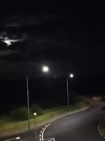 The video shows a nighttime scene with a road in the foreground and a dark expanse in the background, likely a body of water. Two streetlights illuminate the road and the surrounding area. The moon is visible in the upper left portion of the frame, partially obscured by clouds. The lighting suggests it is dark, with the streetlights providing the primary illumination.