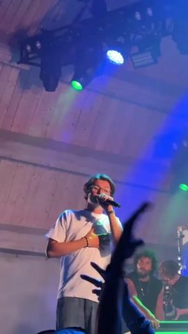 A concert is underway with a performer on stage singing into a microphone, wearing a light-colored t-shirt, jeans, and glasses. They are lit by green and blue stage lights. The audience is visible in the foreground, with some people raising their hands. Another performer is seated on stage to the right, with a beard and wearing a dark t-shirt. The stage has a wooden ceiling. The video then shifts to a wider view of a large outdoor crowd at night, with trees in the background and colored lights illuminating the scene. The camera briefly shows a blurry shot of the performer on stage. The video returns to a shot of the performer on stage with the audience in the foreground.