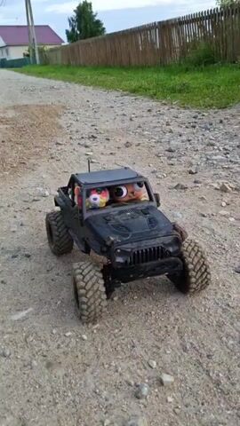 A black remote-controlled Jeep is driving on a gravel road. The Jeep has stuffed animals in the driver's seat and passenger seat. The stuffed animals have large, cartoonish eyes. The road is surrounded by grass and dirt. The video is shaky, as if it was filmed while walking or running.