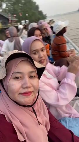 The video opens with a close-up selfie of two young women in hijabs, sitting in a boat. The woman in the foreground, wearing a pink hijab and a maroon shirt, smiles at the camera. Behind her, another woman in a light pink hijab and shirt is visible, holding a water bottle and waving. The boat appears to be moving, as evidenced by the slight blur in the background. Several other people, also wearing hijabs and casual clothing, are seated in the boat behind them. The environment suggests an outdoor setting, possibly a river, with trees and a wooden structure visible along the bank. The lighting indicates it might be late afternoon or early evening. The video continues with a shot of the woman in the foreground covering her mouth and nose, appearing to giggle or react to something. The scene then shifts to a blurry close-up of her face as she laughs.