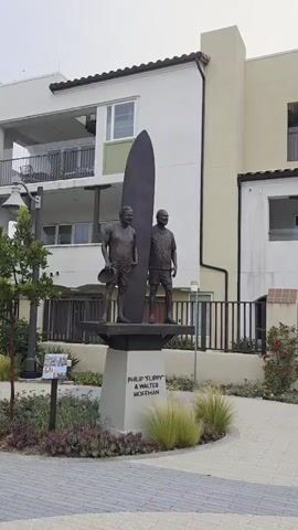 The video shows a panning shot of an outdoor art installation featuring bronze statues of surfers and other individuals, presumably significant figures in surfing history. The statues are mounted on concrete pedestals with inscriptions. One pedestal reads "PHILIP TURPIN & WALTER HOFFMAN". Another reads "JOHN SEVERSON". The statues are arranged in a landscaped area with patterned brick pathways and ornamental grasses. In the background, there are modern residential buildings with white facades, balconies, and windows. The sky is overcast, suggesting a cloudy day.