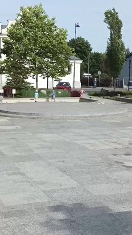 The video shows an outdoor scene with a paved area in the foreground and a slightly elevated circular area with a garden and a building in the background. There are two children playing on the circular area, one wearing a blue shirt and the other a lighter colored shirt. There is also an adult with a stroller and another person walking away from the camera. A grey animal, possibly a dog, is seen in the foreground and on the circular area. A red car is parked in the background near the building. The environment appears to be a public space, possibly a town square, with trees and benches visible. The lighting suggests it is daytime.