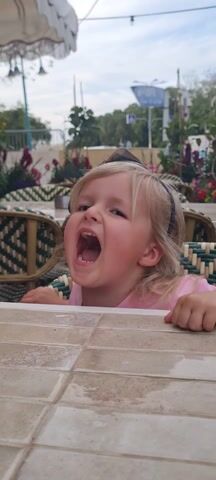 A young girl with blonde hair and a black headband is seated at a table, yelling. She wears a pink shirt. Her hands are on the table's surface. The table is tiled in shades of beige. The chairs have a woven, multicolored pattern.  Behind her, there are flowers in pots and trees. The sky is overcast.