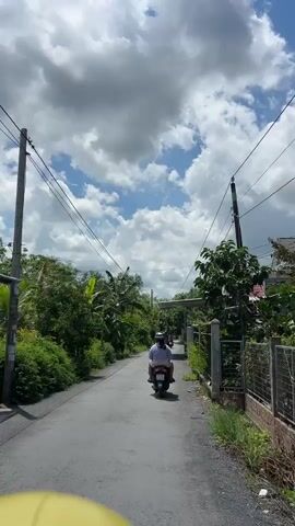 A person rides a scooter down a narrow paved road lined with lush green trees and vegetation. The sky is filled with dramatic white clouds against a bright blue background. Overhead, power lines stretch across the frame. A fence and a small building are visible on the right side of the road. The scooter has a rider wearing a helmet and a white shirt. Another scooter is visible further down the road, also with a rider. The scene appears to be a rural or suburban setting on a sunny day.