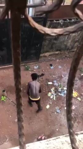 The video, filmed through what appears to be an ornate, brown iron window grill, shows a shirtless man standing on reddish-brown soil. He is facing away from the camera and is wearing dark shorts with a yellow waistband and black sandals. He is surrounded by scattered trash and debris, including what appear to be pieces of paper, plastic bottles, and other unidentifiable items. The man is actively picking up items from the ground and appears to be cleaning the area. Behind him is a dark colored metal door or gate. The lighting is overcast, suggesting an outdoor setting.