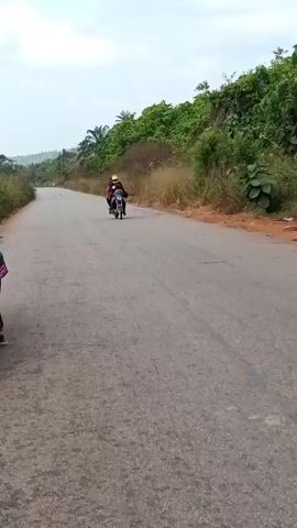 The video shows a grey asphalt road with green vegetation on either side. A motorcycle with three people on it rides down the road. The person driving is wearing a yellow helmet, and one of the passengers is wearing a backpack. A blue car is parked off the side of the road. Someone standing by the open car door watches the motorcycle pass.