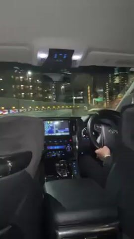 A car drives along a highway at night. The driver sits in the front of the car, wearing a black jacket. From inside the car, high-rise buildings illuminate the cityscape on both sides of the road. The dashboard is visible, including the steering wheel and a navigation screen. A green sign is visible that indicates the direction of travel.