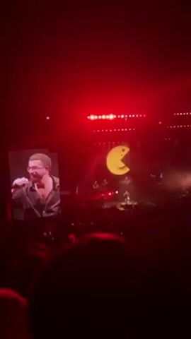 A live music performance is taking place on a large stage. The venue appears to be a large concert hall or arena filled with an audience. A singer is performing center stage, backed by a band. A large video screen behind the stage displays various graphics. A smaller screen to the side of the stage shows a close-up of the singer. The lighting is dramatic, with bright spotlights and colored lights illuminating the stage. The audience can be seen recording the performance with their phones.