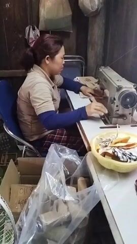 The video shows a woman sitting in a blue chair at a table, sewing on a beige sewing machine. She is wearing a tan shirt with a blue long-sleeved shirt underneath and a plaid skirt. She has her hair pulled back in a ponytail. On the table in front of her are pieces of fabric and a yellow bowl filled with fabric scraps. To her left is a box of items covered in plastic. In the background, there are shelves with various items, including bags and boxes. The room appears to be a workshop or storage area with wooden walls and a slanted ceiling. The lighting is dim. The video is shot from a slightly elevated angle, looking down at the woman.