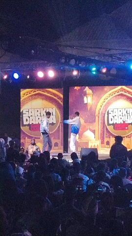 The video shows two men dancing on a stage at an event. They are performing in front of a crowd of people who are seated at tables. The stage has two large screens that say "Sarkin Dariya." The lighting is colorful with spotlights. The crowd is largely silhouetted, but some individuals are visible. Some audience members are recording the performance with their phones.