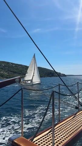 The video shows a serene nautical scene on a sunny day in Luštica, Montenegro. From the perspective of a boat with a wooden slatted deck and a metal railing, another white sailboat is visible in the middle distance, moving across the sparkling blue water. The sailboat has its main sail fully unfurled, catching the sunlight. A gentle wake trails behind the sailboat, indicating its movement.

In the background, a lush green coastline with rolling hills and scattered buildings lines the horizon under a clear blue sky. The sun creates a bright glare in the upper right portion of the frame. The water is a deep blue with ripples and small waves, reflecting the sunlight. The scene suggests a pleasant day for boating or sailing. No discernible people or text are visible in the video.