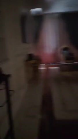 The video pans across a dimly lit room. In the foreground, a dark valet stand with a hanger is visible. Behind it, a wall features built-in shelves with decorative items, including what appear to be circular objects and a cylindrical vase. Further back, a wall-mounted television is mounted on a paneled wall with ornate gold trim. To the right, a console table with a gold ornate base is positioned beneath the TV. Beyond that, the scene opens to a living area with a plush armchair, a coffee table, and draped curtains in shades of pink and grey.