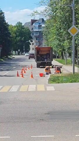 The video shows a street scene in Lytkarino, Russia, on a sunny day. The main subjects are two construction workers performing roadwork. They are kneeling near an open truck, which appears to be carrying materials and tools for the job. Orange traffic cones are placed on the road, marking the work area and directing traffic. A crosswalk is visible in the foreground, with yellow and white stripes.

In the background, a multi-story building with a distinctive architectural style is visible, along with mature trees lining the street. Several cars are driving on the road, including a silver SUV and a red car, indicating that traffic is flowing despite the roadwork. A yellow diamond-shaped traffic sign is mounted on a pole on the right side of the frame. The sky is clear and blue with some scattered clouds, suggesting fair weather. The overall atmosphere seems to be one of normal urban activity with ongoing public works. No text is clearly legible in the video.