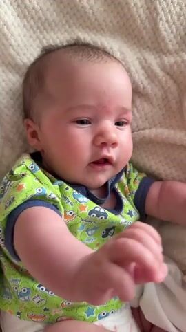 A close-up, eye-level shot shows a baby lying on a textured white blanket. The baby is wearing a green short-sleeved onesie with a repeating pattern of cars, stars, and other graphics. The baby's head is turned to the right, and its expression is calm. The baby's arms are extended forward. The lighting is soft and diffused.