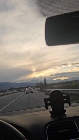 A static shot from inside a car driving on a multi-lane road at sunset or sunrise. Other vehicles are on the road. Mountains are visible in the distance under a cloudy sky. A phone holder is attached to the dashboard. A rearview mirror is visible. The steering wheel is visible on the left. A section of the car's sun visor with a dot pattern is also visible. The sound of music is audible.