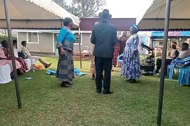 The video shows an outdoor gathering under tents. In the foreground, two women in traditional attire and a man in a suit and hat are interacting. The woman on the left wears a blue top and a patterned skirt. The woman on the right wears a blue headwrap and a blue and white patterned dress. The man is facing away from the camera. To the left, a group of people are seated on chairs under a tent. To the right, another group is seated, and there is some audio equipment visible. The ground is covered in grass, and there are some scattered fabric pieces. The scene appears to be a social event, possibly a ceremony or celebration.