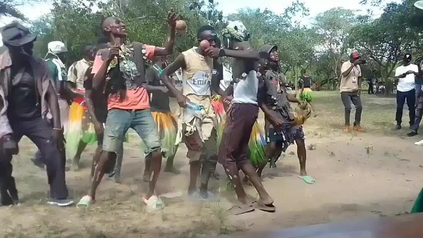 Sure, here is a brief description of the video:

The video shows a group of people dancing in a dirt field in what appears to be an outdoor community event. There are nine people in the group who are dancing. Some members of the group are holding coconuts. The group appears to be enjoying themselves. There are other people standing around the group watching the dance, including a person in a white shirt who is taking a picture or video. The environment is wooded, and there are other people seated in the background.