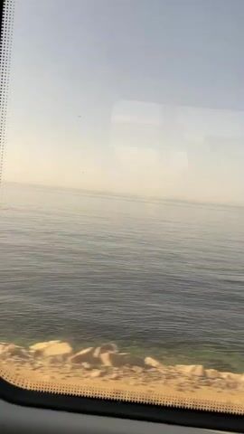 The video shows a view from a moving vehicle, likely a train, looking out a window at a calm body of water and a clear sky. The water appears to be shallow near the shore, with visible greenish hues indicating the seabed or aquatic plants. The shoreline is sandy and rocky. A tall, metal pole with a ladder and signal lights is visible on the right side of the frame at one point. The sun is casting a warm glow, creating a hazy atmosphere. The reflection of the interior of the vehicle is visible on the window.
