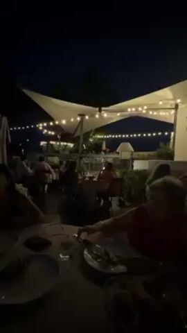 Here is a description of the video:

The video captures a nighttime scene at an outdoor restaurant. People are seated at various tables, enjoying their meals under the illumination of strung lights and overhead canopies. The tables are set with plates, glasses, and some food items. The atmosphere appears relaxed and social, with diners engaged in conversations. The background reveals hints of surrounding greenery and other light sources, contributing to the ambiance of an outdoor dining experience.