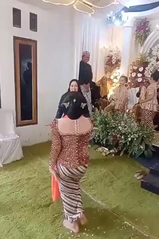 In a room decorated with white drapes and floral arrangements, a wedding ceremony is taking place. Several individuals, some in traditional attire including sarongs and kebaya, are gathered. The main focus is on a woman in a brown sequined kebaya and a striped skirt, who is dancing. She is barefoot and has her hands raised in a dancing motion. She is interacting with another woman in a black headscarf and a similar striped skirt, who is also in a traditional outfit. In the background, two men in traditional attire stand facing the audience, and several children are also present. The room has a green carpeted floor, and a large window with a dark reflection is visible on the left wall.