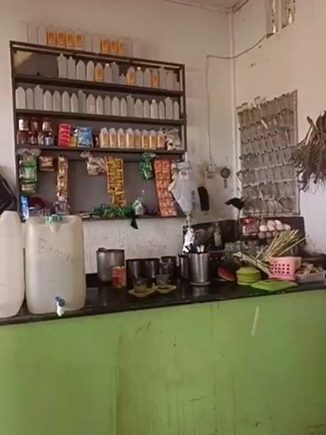 This is a close-up, medium shot video showing a counter with various items on it. The counter is green and appears to be made of wood. On the counter are two large white plastic containers with blue spigots. The container on the left has "Bitcoin" written on it in black marker, and the container on the right also has "Bitcoin" written on it. Behind the counter is a black shelf unit filled with bottles and packaged goods. The shelves are packed with items of various sizes and colors. On the far right of the counter, there are some green leafy vegetables and a pink basket. The background is a plain white wall. The lighting is overhead and appears to be artificial. The video is shaky and the focus is not always sharp. There are no people visible in the video.