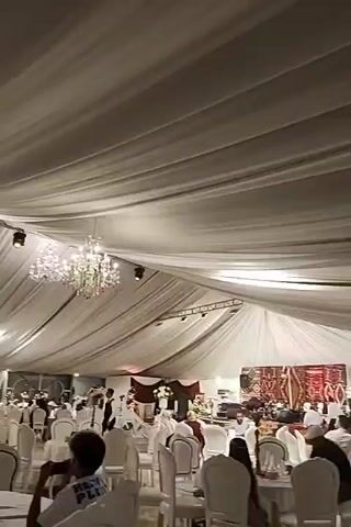 This is a video clip of an indoor event, likely a wedding or formal gathering. The ceiling is draped with white fabric, creating a tent-like atmosphere, with a large, ornate chandelier hanging from the center. White chairs are arranged around tables, indicating a dining or seating area. Several people are visible, some seated and others standing. In the background, there appears to be a stage or decorated area with a screen or backdrop. The lighting is dim, with spotlights illuminating certain areas. The overall impression is one of elegance and festivity.