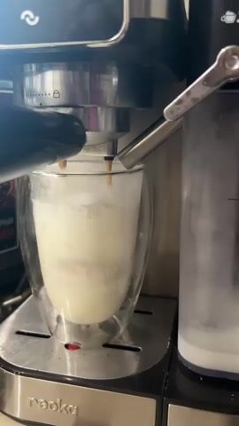 A close-up shot shows a coffee maker dispensing espresso into a double-walled glass mug filled with milk. The espresso streams down from the coffee maker, creating layers of brown coffee and white milk in the mug. The coffee maker has a chrome finish and a black handle. The mug is clear glass, allowing the layers of liquid to be visible. The scene appears to be in a kitchen or coffee shop.