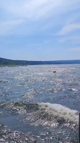 The video shows a body of water with waves. The water is blue and reflects the sky. There is a green hill in the background. There is also a red buoy in the distance. The camera is moving, giving the impression of being on a boat.